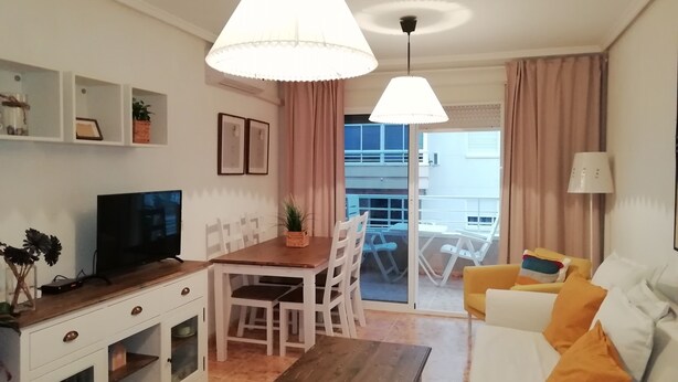 Una sala de estar moderna con una mesa de centro de madera, un sofá blanco y un comedor con una mesa y sillas de madera. Hay un televisor sobre un mueble bajo, y un balcón con vistas a edificios.
