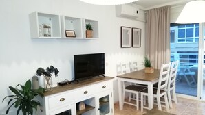 Un salón moderno con televisor de pantalla plana, muebles blancos, una mesa de comedor con sillas y un ventanal con vista a un edificio.