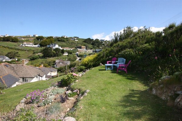 A hillside garden with a seating area featuring two chairs and a table, surrounded by lush greenery and flowering plants.