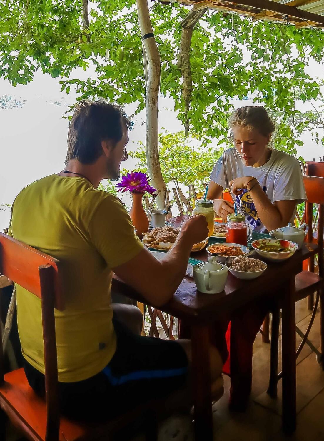 Deux personnes sont assises à une table en plein air, devant divers plats et boissons, et sont entourées de verdure.