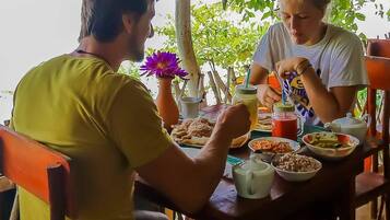Deux personnes sont assises à une table en plein air, devant divers plats et boissons, et sont entourées de verdure.