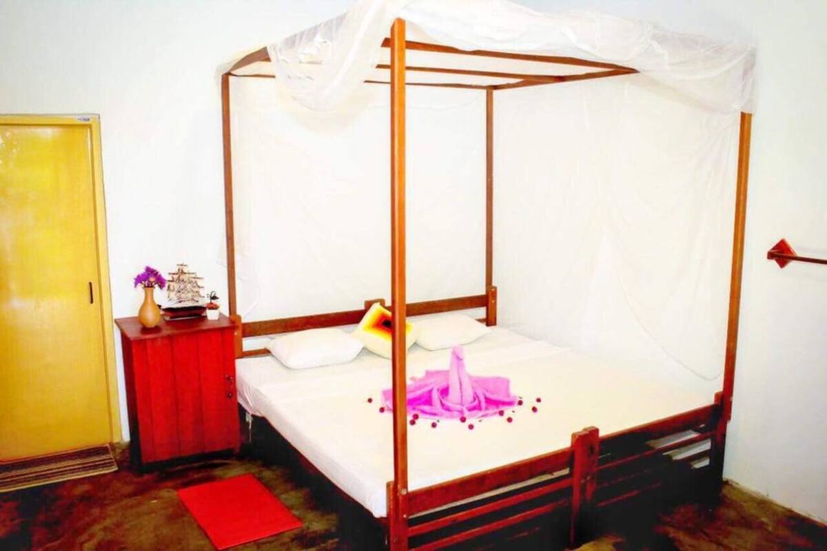 Un lit à baldaquin, une armoire en bois, un tapis rouge et une petite table sur laquelle sont posés un vase et une maquette de bateau.