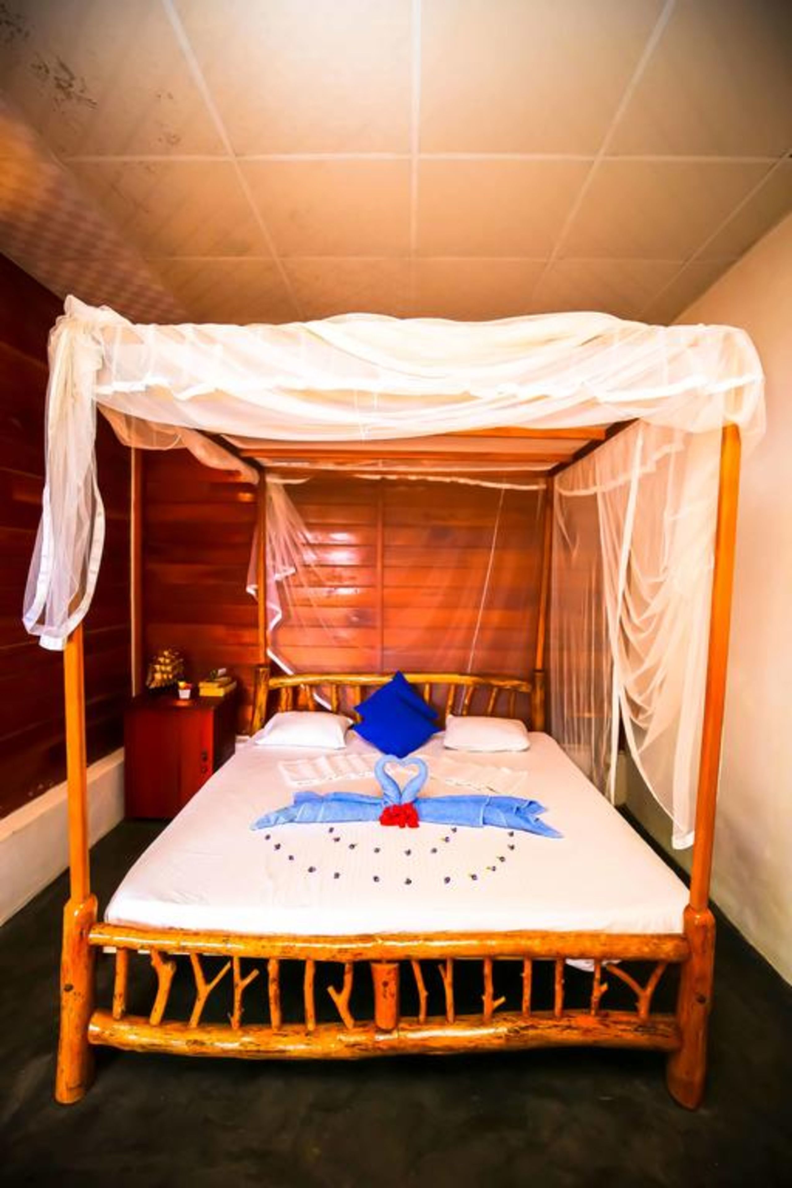 A wooden four-poster bed with a white canopy, a blue pillow, and a red heart-shaped decoration on the bedspread.