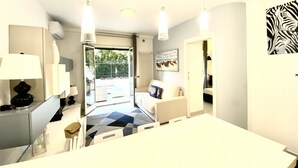 A modern kitchen with a white countertop, a stove, and a sink. There is a dining area with a white table and chairs. The room has a large window and a view of the outdoors.