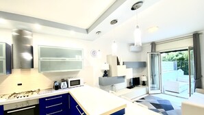 A modern kitchen with blue cabinets, a white countertop, and a built-in oven. There is a dining area with a white table and chairs, and a large window offering a view of the outdoors.