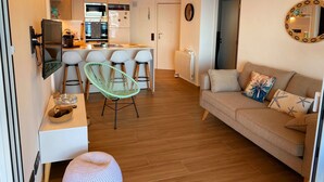 A modern living room with a sofa, a dining area with bar stools, a TV mounted on the wall, and a kitchen with a built-in oven and microwave. - Stunning beachfront penthouse (Calonge)