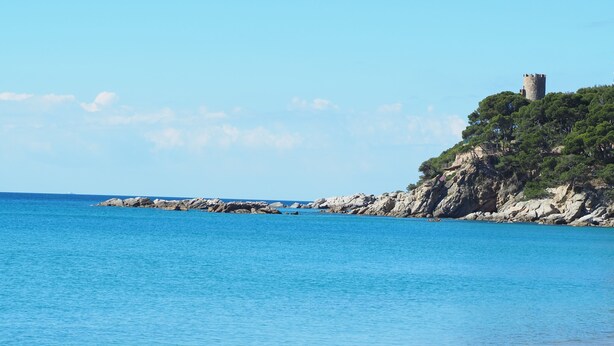 A coastal landscape with a sandy beach, calm blue waters, rocky outcrops, and a tower on a hill. - Stunning beachfront penthouse (Calonge)