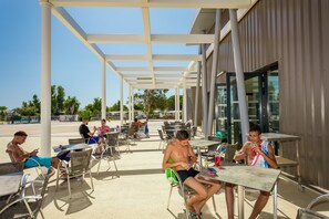 An outdoor cafe with metal chairs and tables, a covered walkway, and a building with large windows. - Camping des Rives des Corbieres (Leucate)