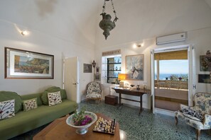 Una habitación con un sofá verde, un tablero de ajedrez, una mesa con una lámpara y vista al mar a través de una puerta.