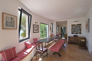 Un comedor con una mesa larga, bancos y sillas. Hay obras de arte enmarcadas en las paredes y vistas al exterior con vegetación.