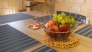 Uma área de refeições com uma mesa de madeira, um placemat tecido e uma fruteira com maçãs e uvas. Uma escada em espiral é visível ao fundo.