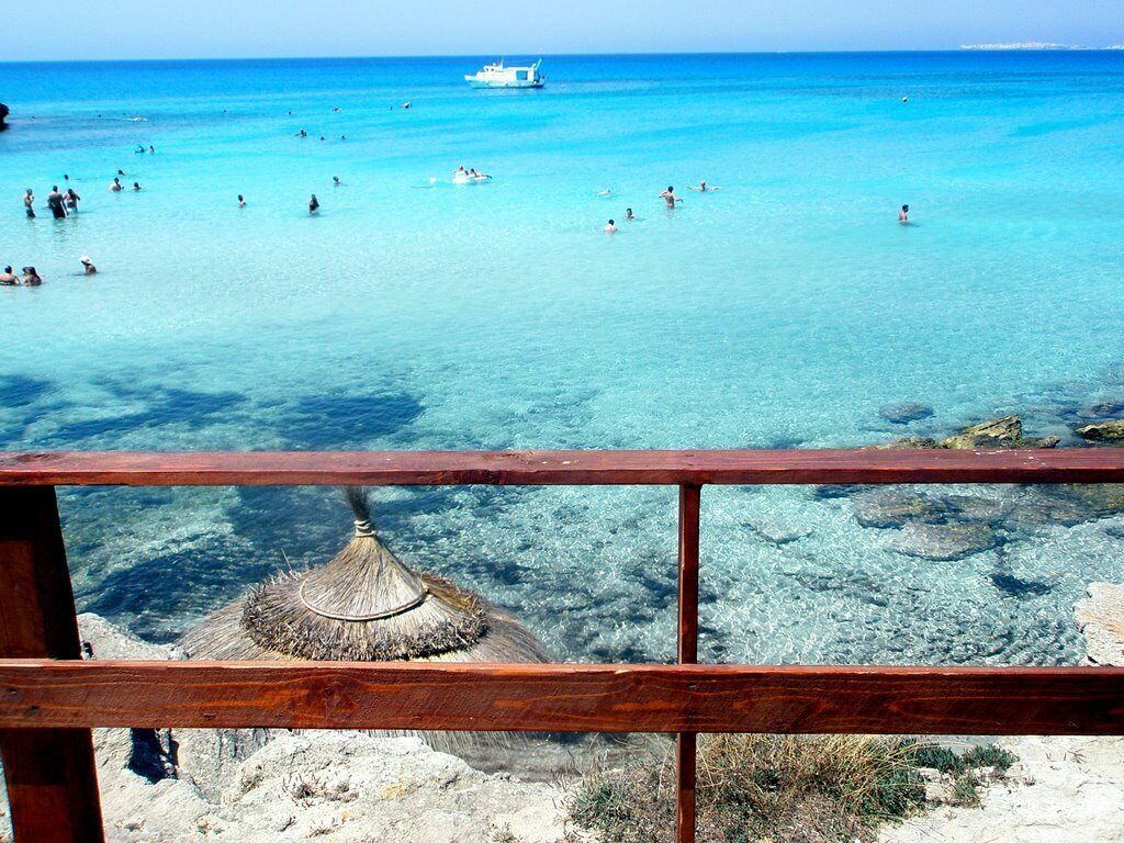 Ein Strand mit klarem türkisblauem Wasser, einem hölzernen Geländer und einer reetgedeckten Hütte im Vordergrund.