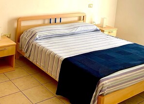 A single bed with a striped bedspread and a dark blue blanket, a wooden nightstand with a lamp, and a small drawer unit.