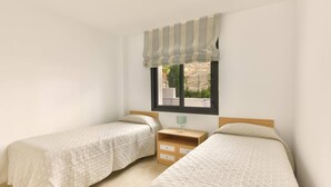 Een slaapkamer met twee bedden, een raam met een gestreept gordijn, een nachtkastje met een lamp en een klein dressoir.
