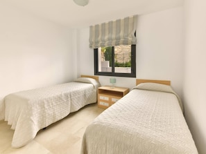 Un dormitorio con dos camas, una ventana con un visillo a rayas, una mesita de noche con lámpara y una cómoda pequeña.