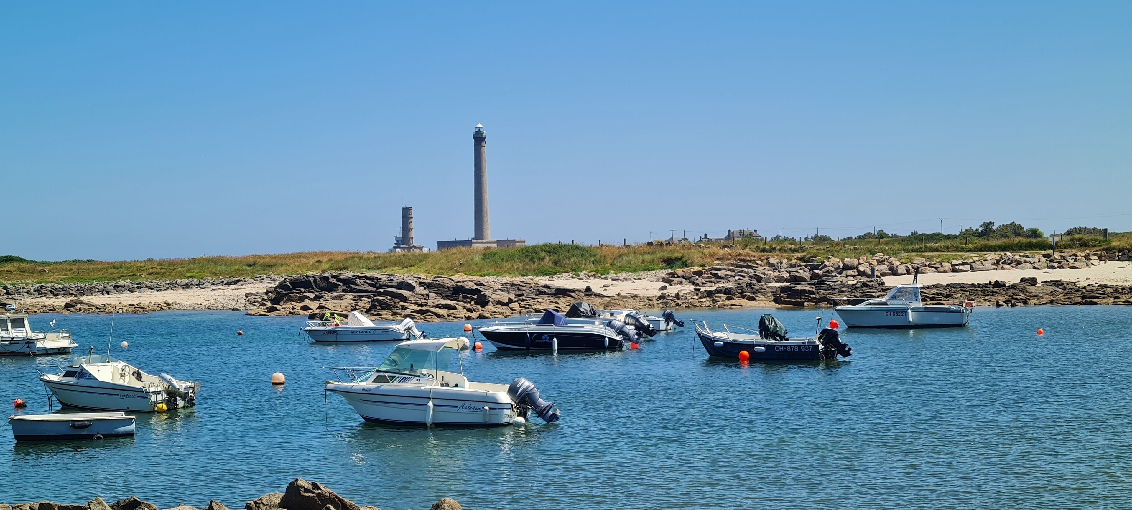 A marina with several boats docked, a lighthouse in the background, and rocky terrain.