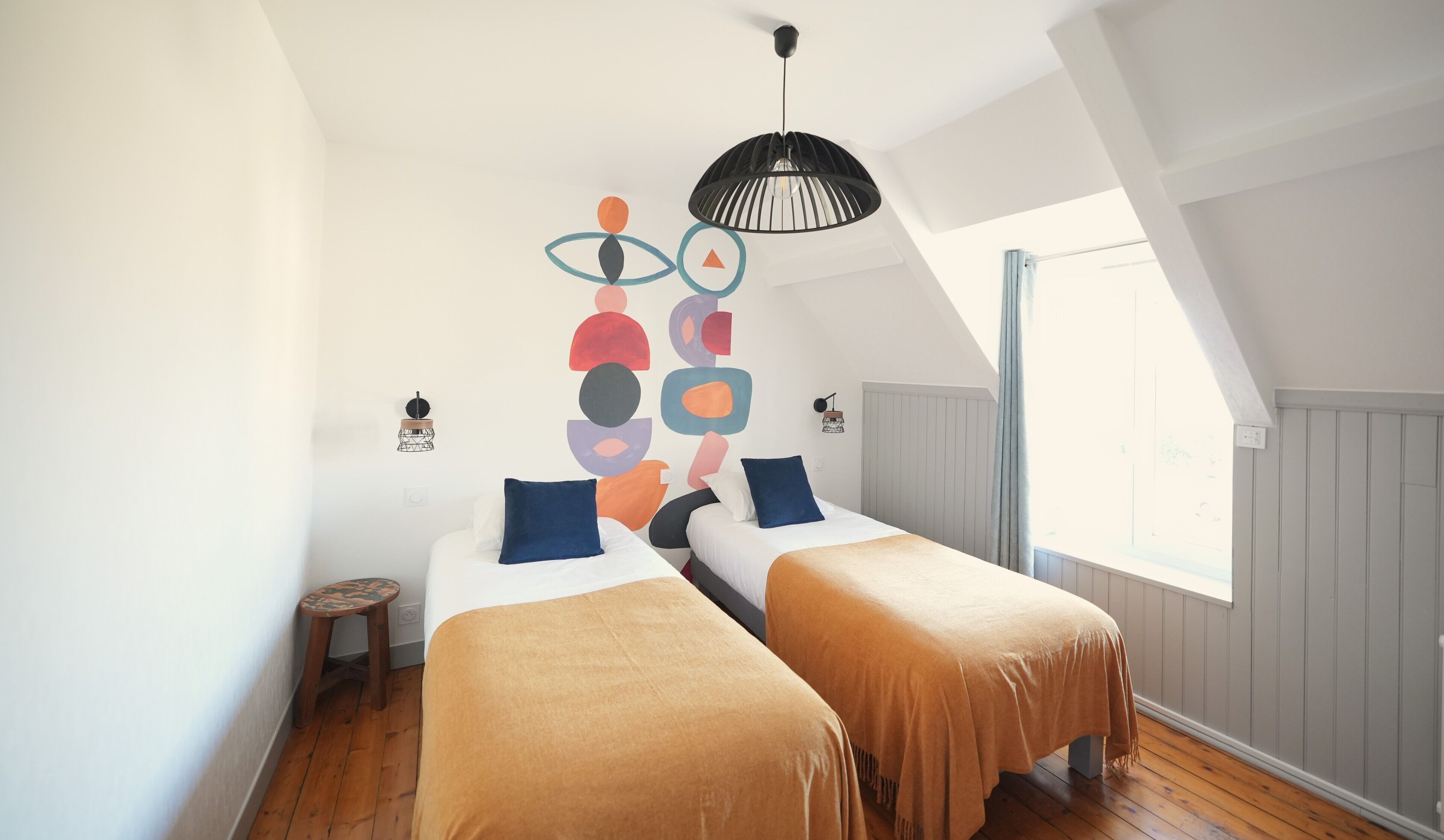 A bedroom with two beds, a small stool, a ceiling light, and a window.