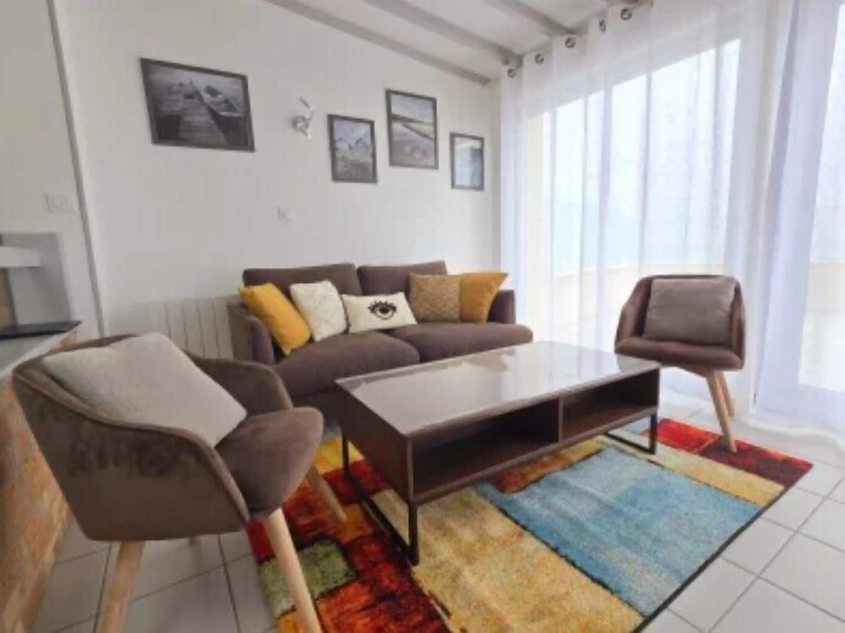 Un salon moderne avec un canapé marron, une table basse et deux fauteuils. Trois tableaux encadrés ornent le mur et un tapis coloré recouvre le sol.