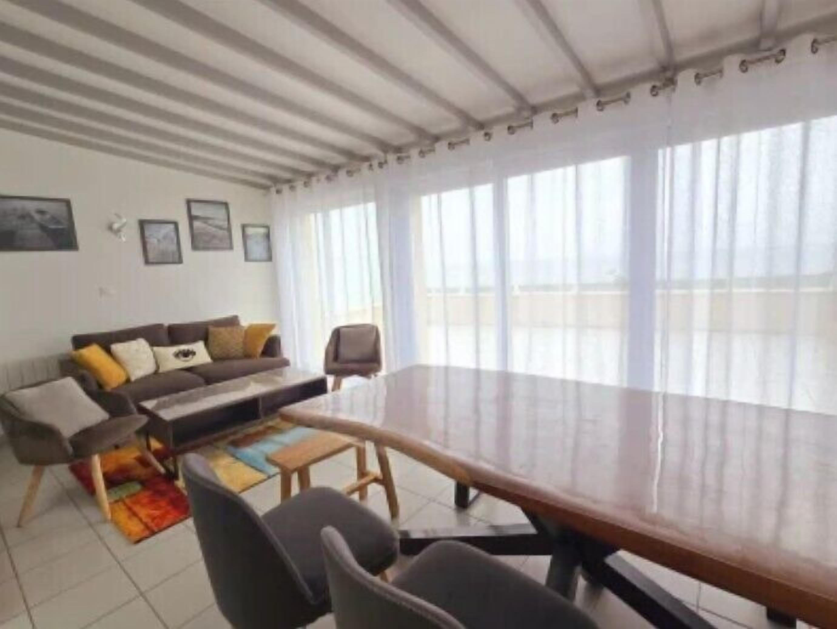 A spacious living room with a large wooden table, a sofa with cushions, and a view through large windows.