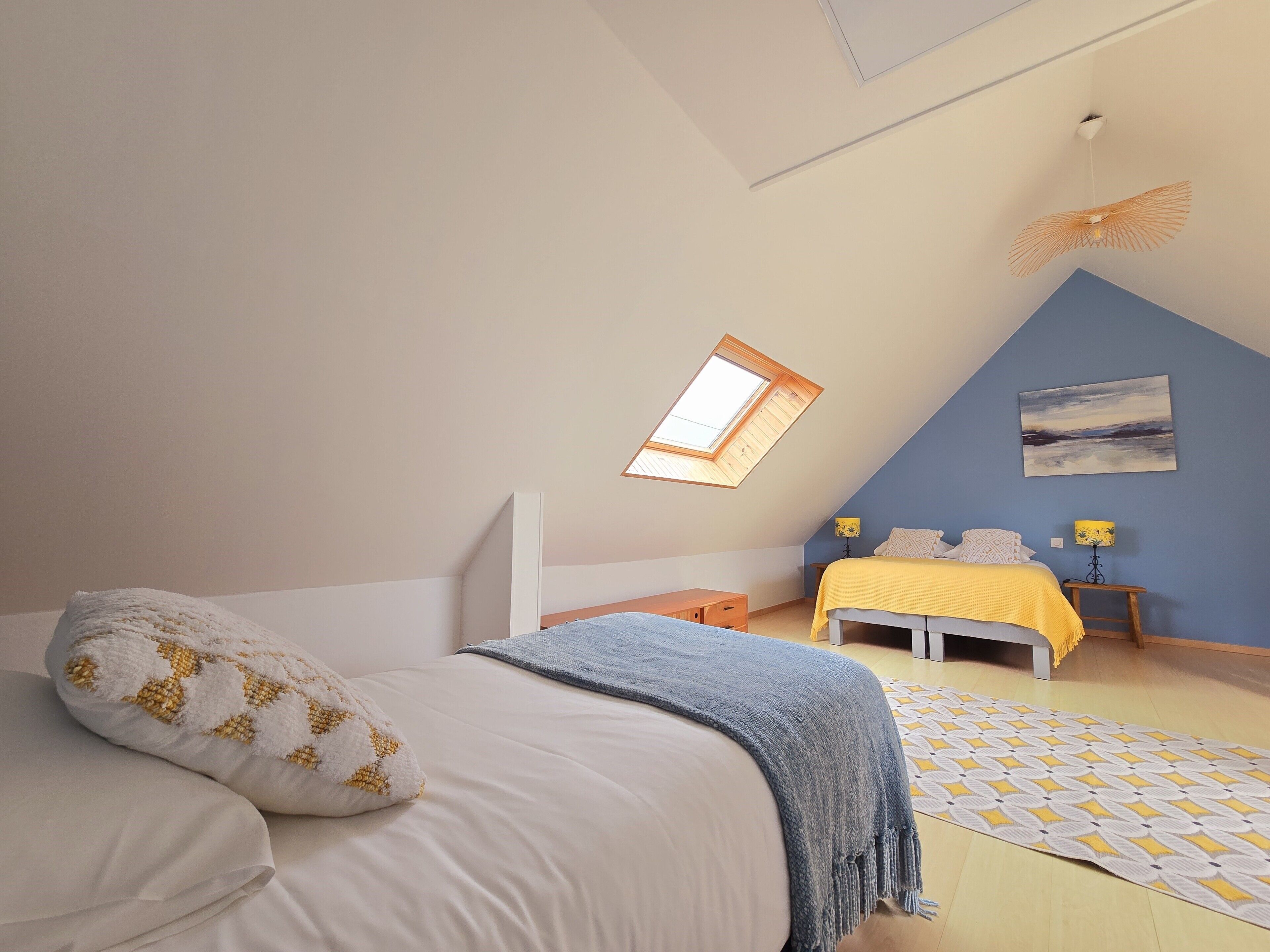 A bedroom with a sloped ceiling, a skylight, a bed with a blue blanket, a bedside table with a lamp, and a painting on the wall.