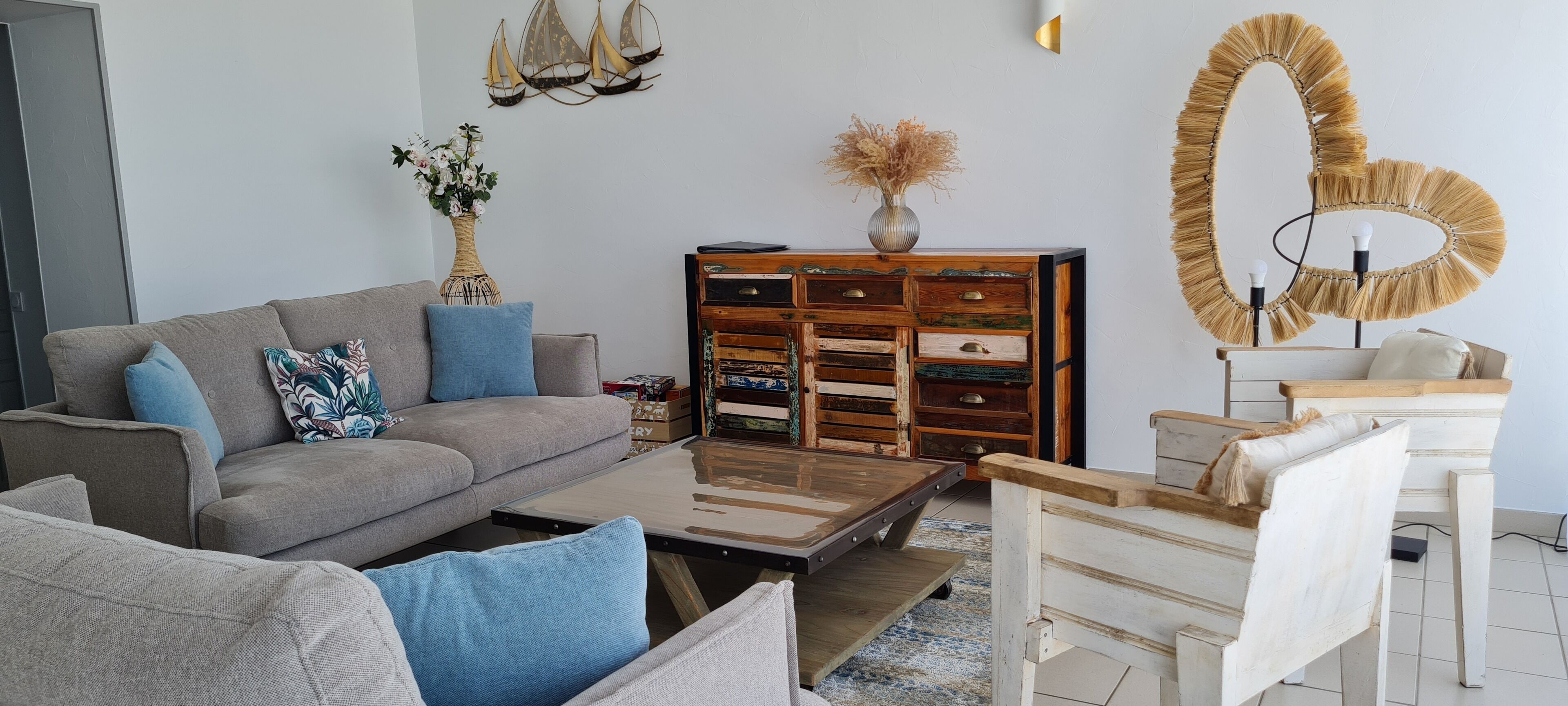 Een woonkamer met een grijze bank, blauwe kussens, een bijzettafeltje, een houten kast en een decoratieve spiegel.