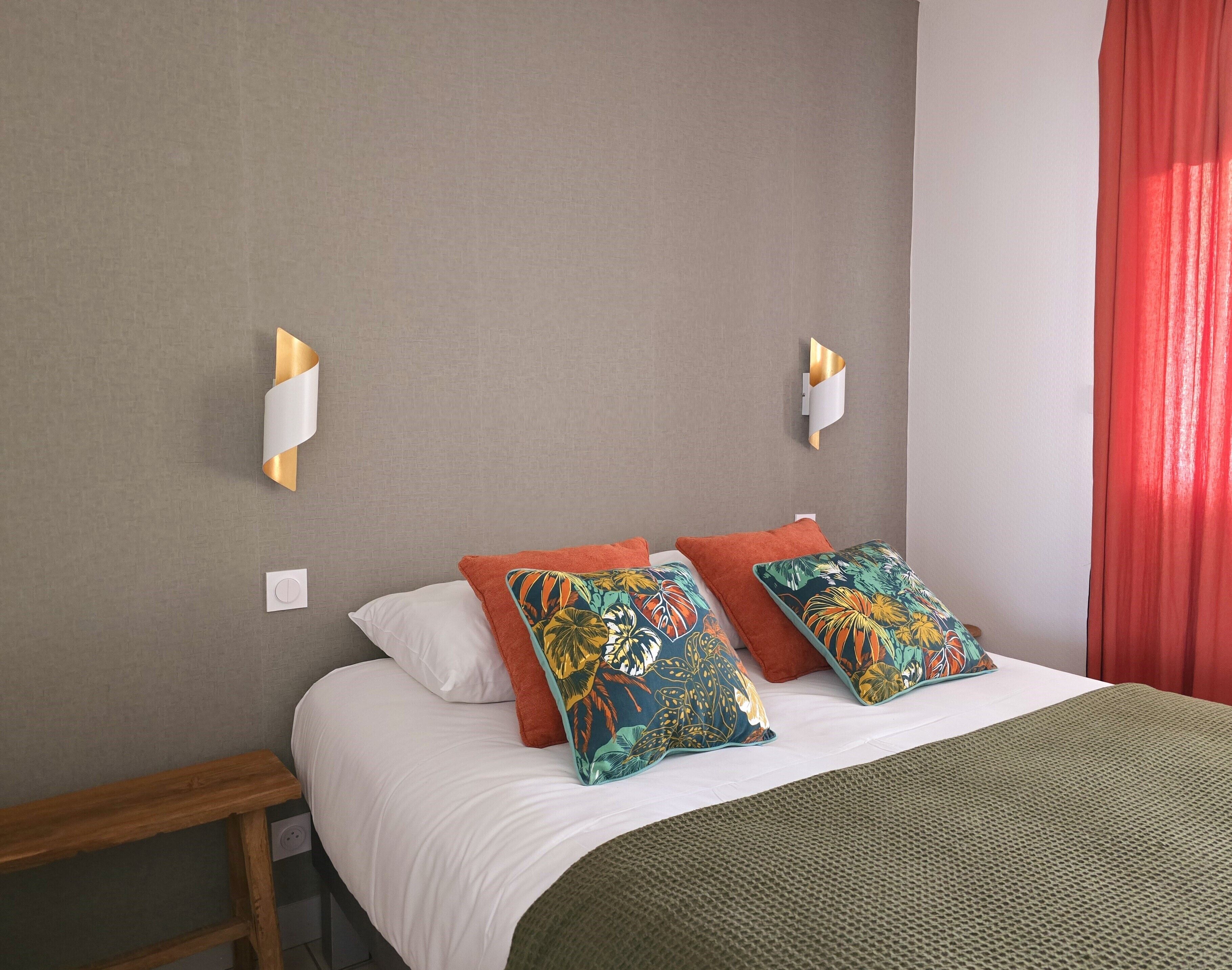 Uma cama bem arrumada com lençóis brancos e duas almofadas coloridas, uma mesinha de cabeceira de madeira e um cobertor verde com textura.
