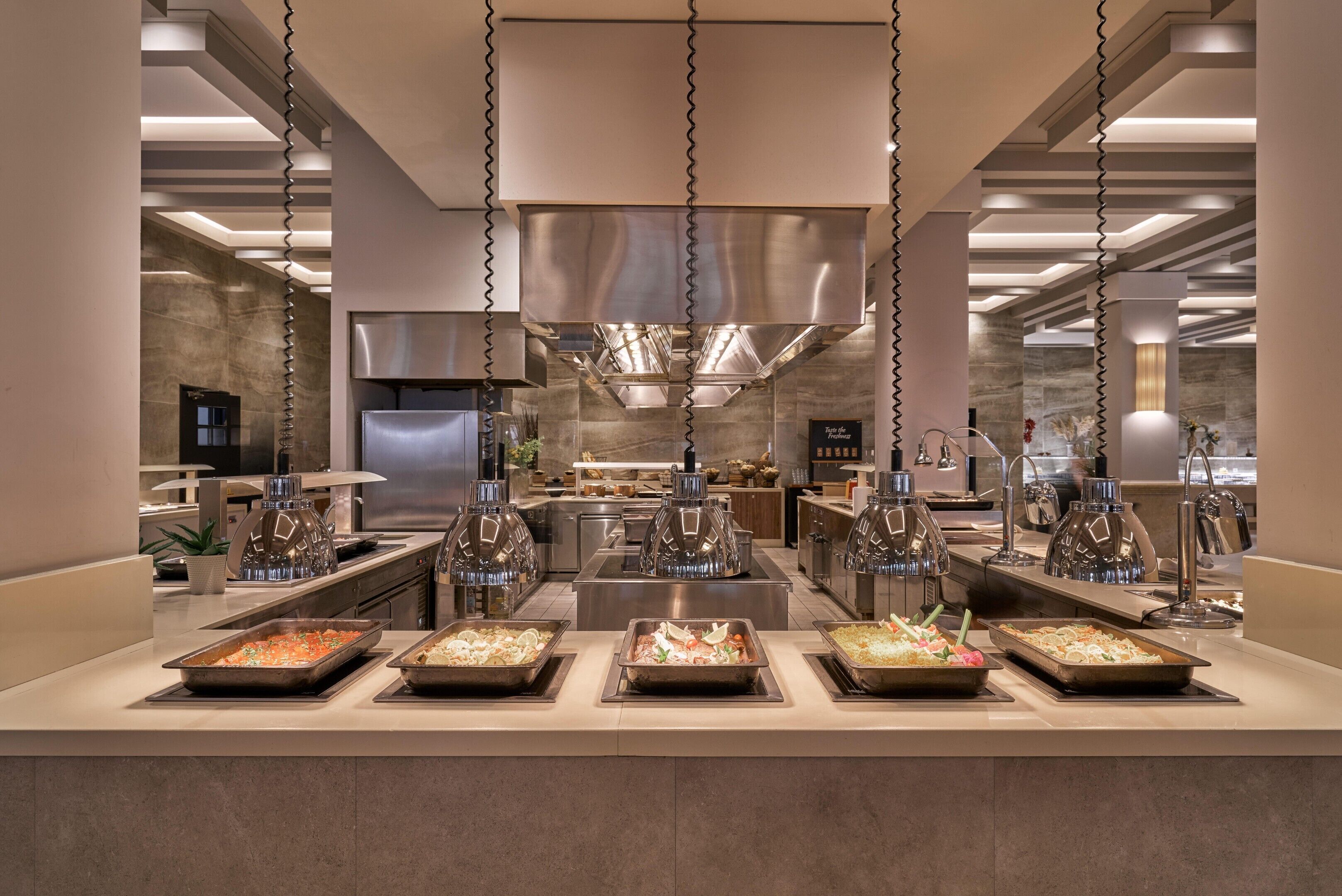 Montaje de bufé con diferentes estaciones de comida y una cocina moderna al fondo.