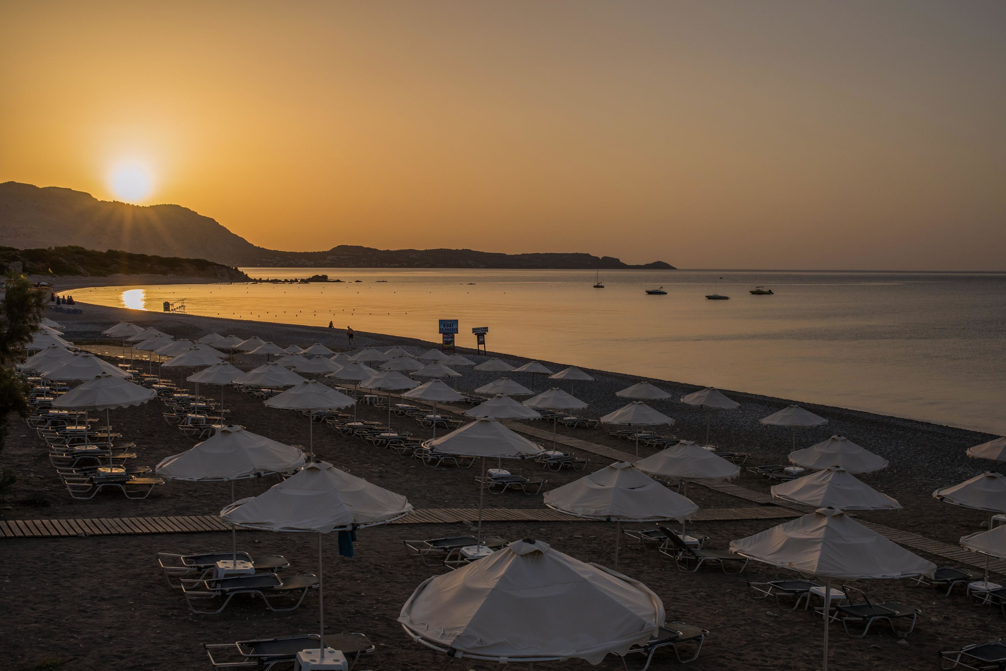 Uma praia com fileiras de guarda-sóis e espreguiçadeiras, mar calmo e um pôr do sol ao fundo.