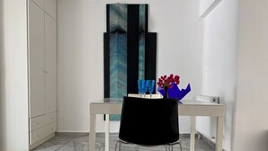 Et værelse med et hvidt skab, et hvidt bord med en sort stol, et stort abstrakt maleri og en vase med røde blomster.