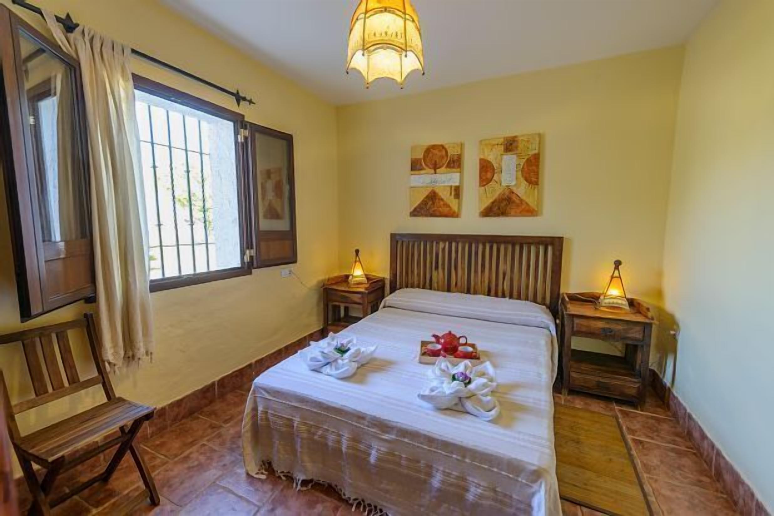 Un cuarto de hotel con una cama grande, dos mesitas de noche con lámparas, un banco de madera y obras de arte en las paredes.
