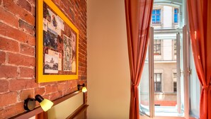 Una habitación con pared de ladrillo, un collage enmarcado, una cama con almohadas, un radiador y una ventana con cortinas.