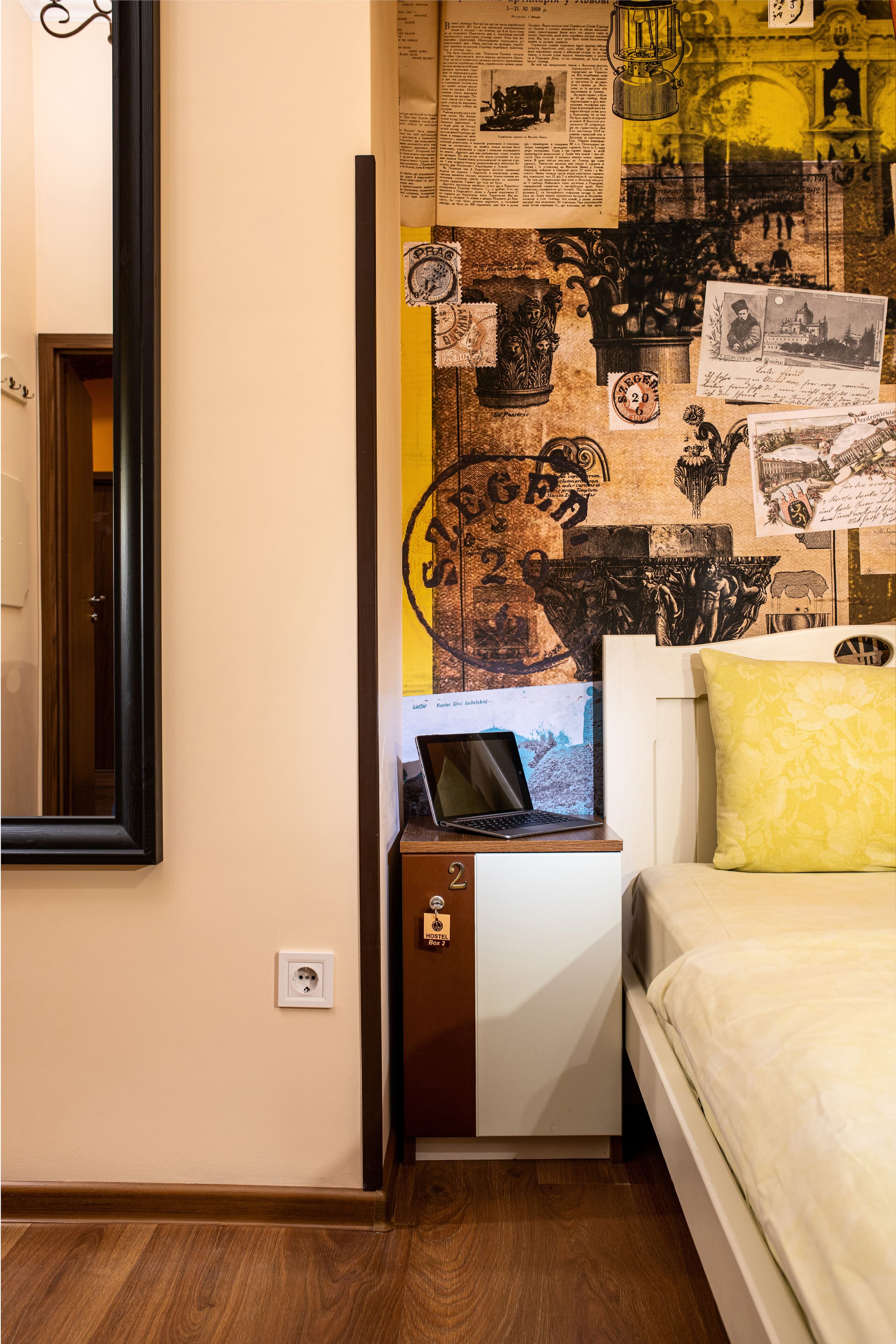 Une chambre à coucher avec un lit, une table de chevet sur laquelle se trouve un ordinateur portable, un mur décoré d’anciennes affiches et une horloge.