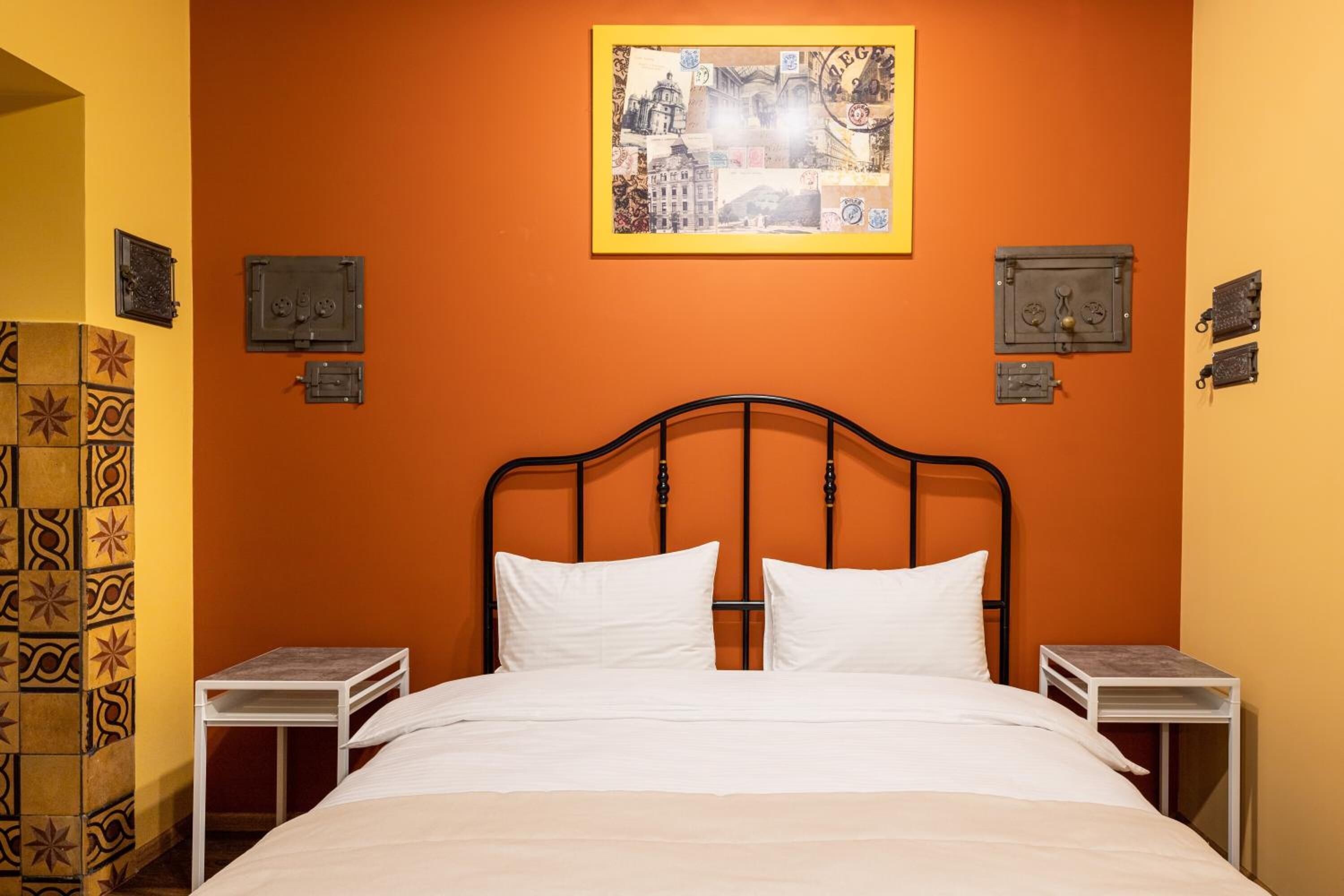 A neatly made bed with white linens in a room with orange walls, a framed cityscape artwork above the bed, and two bedside tables with small safes.