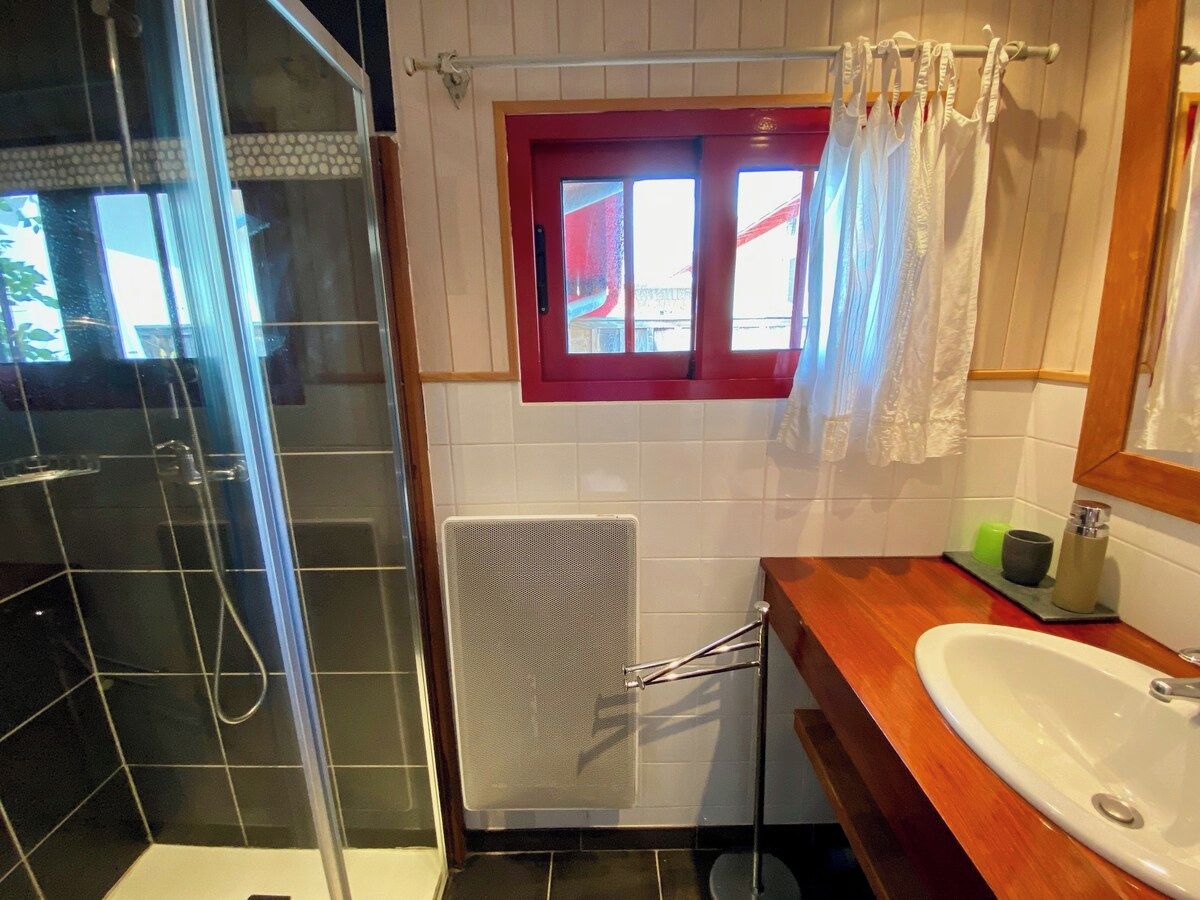 Ein Badezimmer mit Dusche, einem Fenster mit rotem Rahmen, einem Handtuchhalter und einem Waschbecken mit Spiegel.