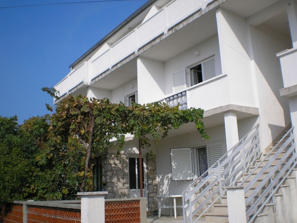 A white multi-story building with a staircase leading to the entrance, a small outdoor seating area, and a tree with green leaves in front.