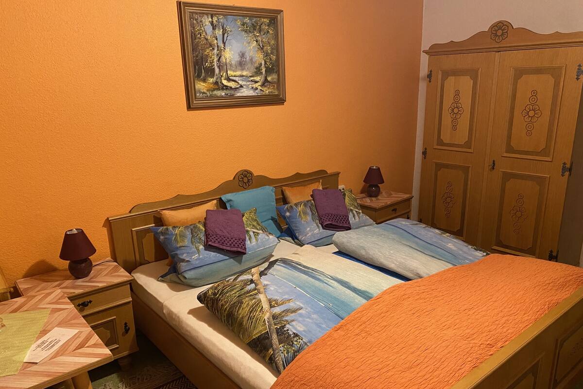 A bedroom with a wooden bed, multiple pillows, a nightstand with a lamp, a wooden wardrobe, and a painting on the wall.