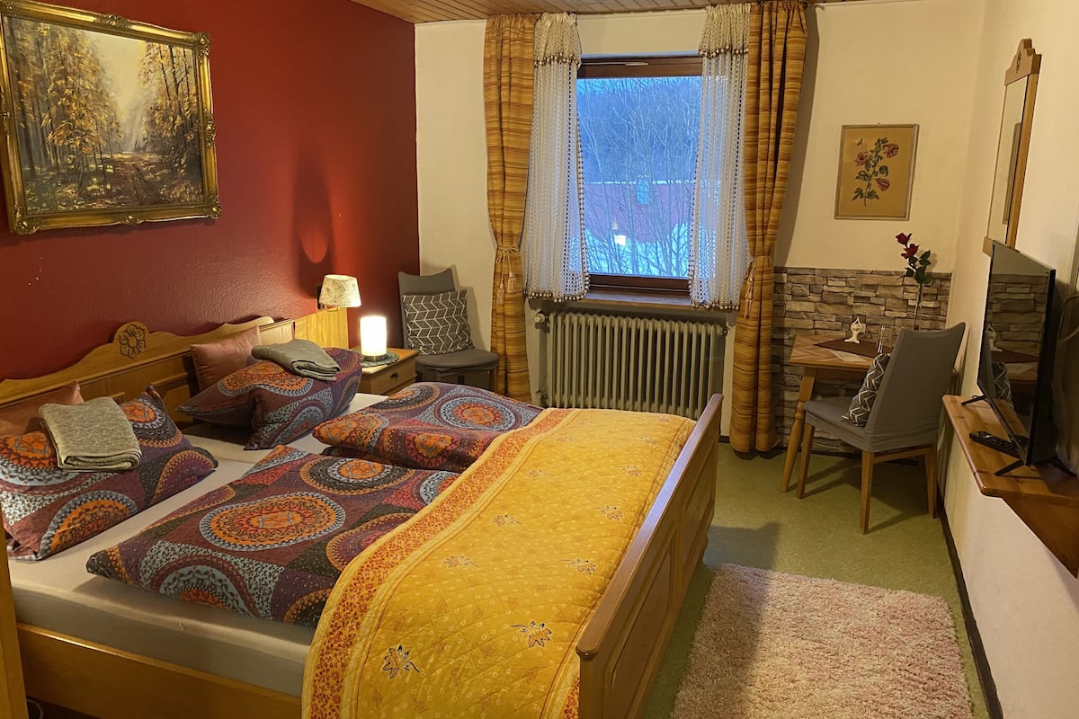 A cozy bedroom with a bed, a chair, a table, a painting, and a window with curtains.