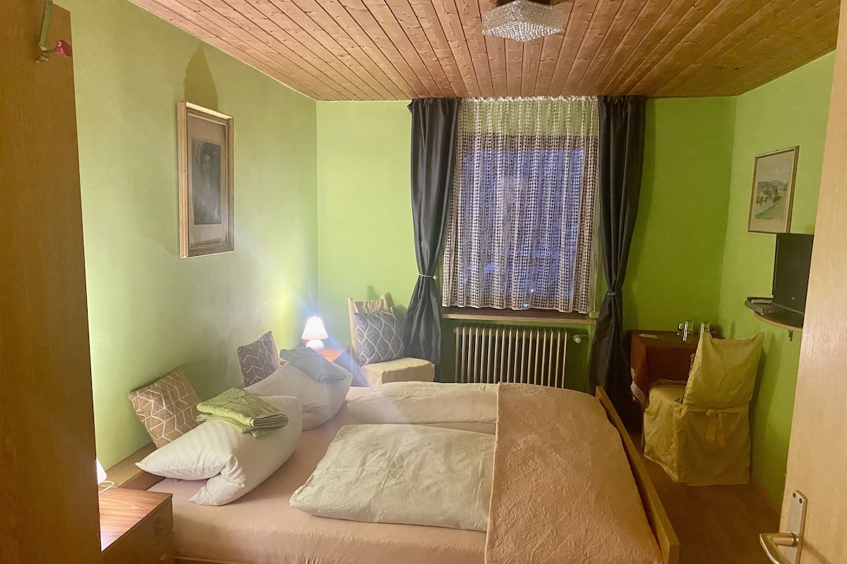 A room with a bed, a nightstand, a lamp, a window with curtains, a radiator, and a picture on the wall.