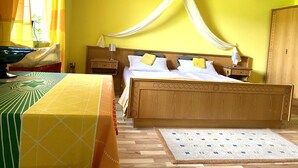 A room with a wooden bed, a nightstand, a bedside table, a table with a patterned tablecloth, and a window with curtains. - Pension Waldblick (Spiegelau)