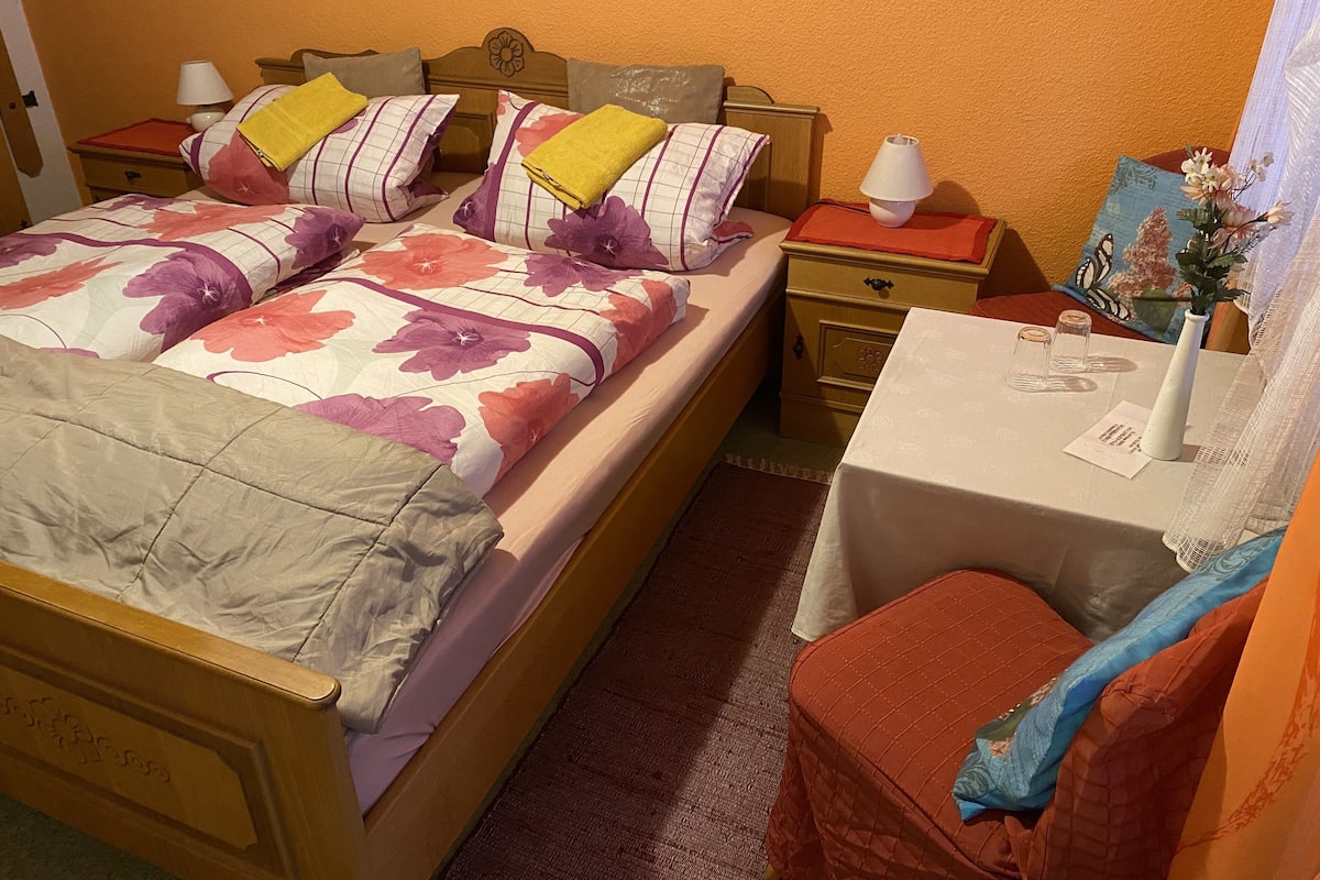 A neatly arranged bedroom with a bed, bedside tables, a small table with a vase, and a chair.
