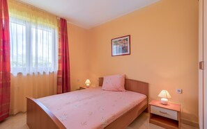 Un dormitorio con cama, mesita de noche con lámpara, ventana con cortinas e imagen colgada en la pared.