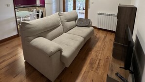 A living room with a beige sofa, wooden flooring, a TV stand, and a dining area in the background. - Apartment for 4 people (Usurbil)