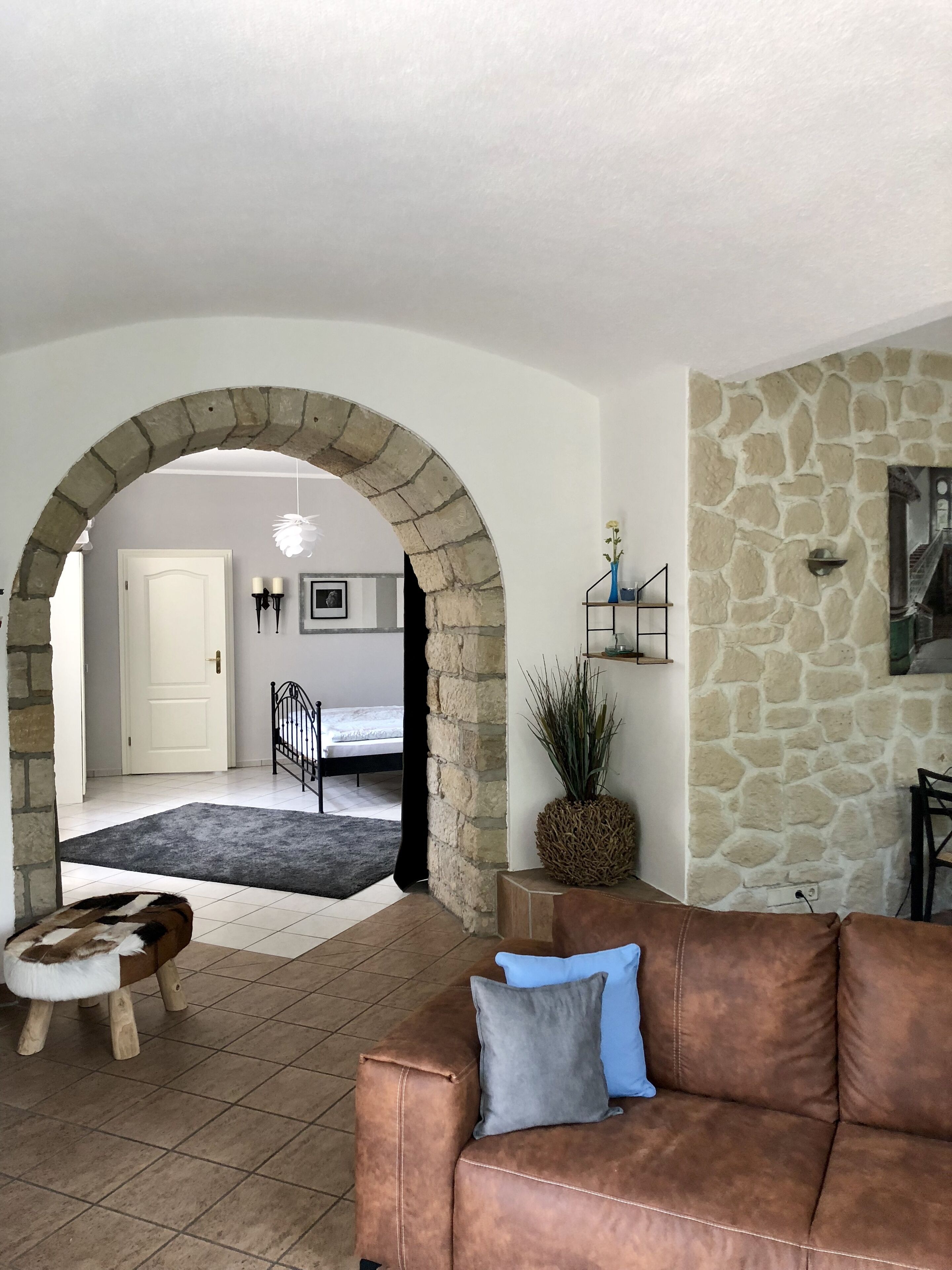 Ein Wohnzimmer mit Steinwand, einem braunen Ledersofa, einem kleinen Holzschemel und einem schwarzen Metallgitter, das in ein Schlafzimmer führt.