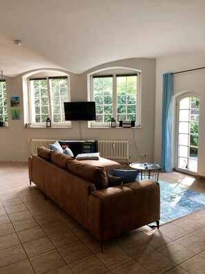 A living room with a brown sofa, a flat-screen TV, a radiator, and large windows with curtains. - Vacation apartment in the Villa Graf von Pfeil on the banks of the Elbe (Neustadt)