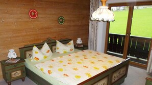 A bedroom with a wooden bed, floral bedding, a nightstand with a lamp, and a view of a green field through a window.