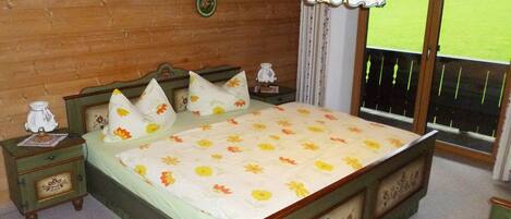 Un dormitorio con cama de madera, ropa de cama floral, una mesita de noche con lámpara y vistas a un campo verde a través de una ventana.