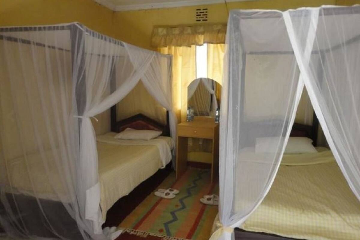 Een kamer met twee bedden, elk voorzien van een klamboe. Er staat een klein tafeltje met een spiegel en een lamp. De muren zijn geel en er ligt een kleurrijk vloerkleed op de vloer.