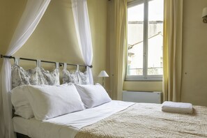 Cama bem arrumada com lençóis brancos e colcha bege, luminária de cabeceira e janela com cortinas.