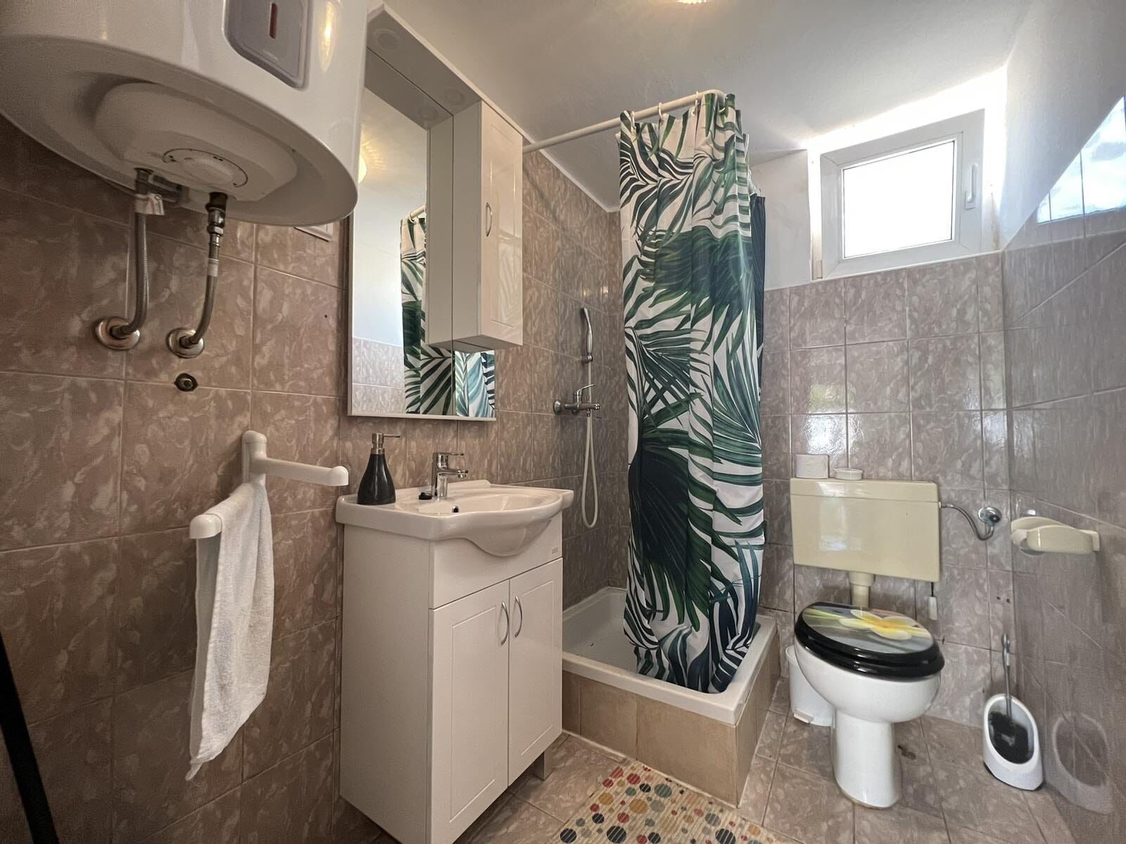 Une salle de bain moderne avec un meuble-lavabo blanc, une douche avec un rideau à motifs de feuilles vertes, des toilettes et une petite fenêtre.