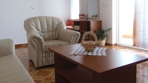 Una sala de estar con un sillón color beige, una mesa de centro de madera y un escritorio con una silla. Se ve una ventana con cortinas y una planta en el alféizar.